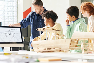 A small group of grade eight students are directed to study blue prints of an airplane on a computer by their teacher, on the desk beside them are exact model replicas