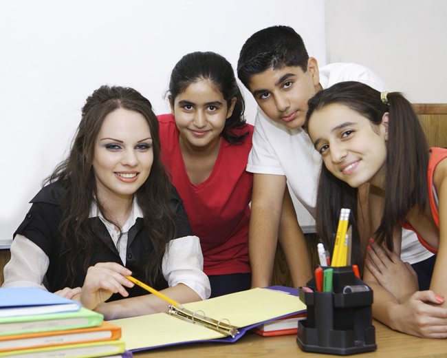 Teacher at her desk with three students there for group photo Open Gallery