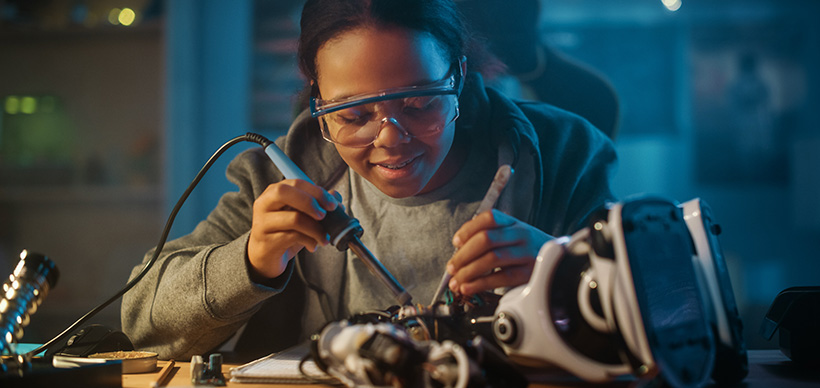 A safety goggled female student leans over her desk as she solders robotic circuitry