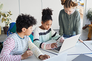 Three grade seven students are seated at a desk, in the middle is a laptop, all three happily coordinate the sharing of the computer