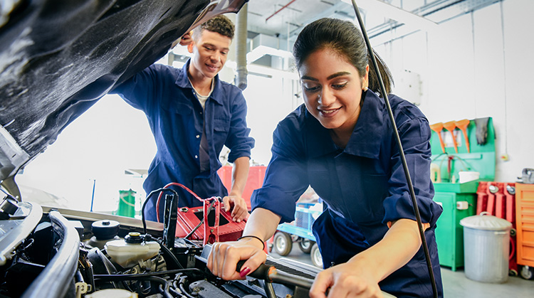 Two high school students, wearing one piece blue overalls have the hood of a car open and they are working on the motor with a ratchet