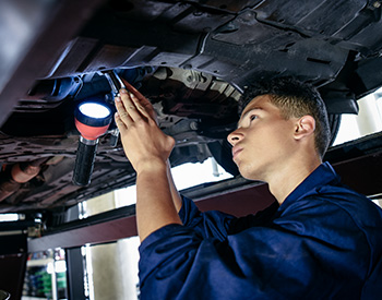 A high school auto mechanic student explores the undercarriage of a hoisted vehicle with wrench and flashlight