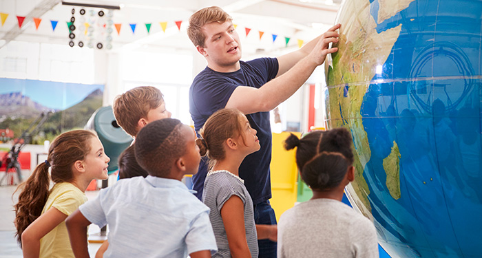 In a bright and spacious classroom, a young teacher directs the attention of his eager cluster of middle school students to a specific location on the comically large globe before them