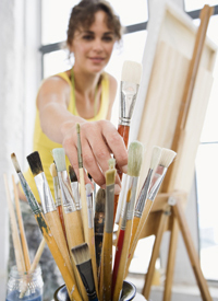 Artist reaching for a paintbrush just beyond the easel and canvas 