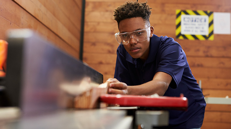 A young safety goggled high school student carefully pushes wood through a table saw